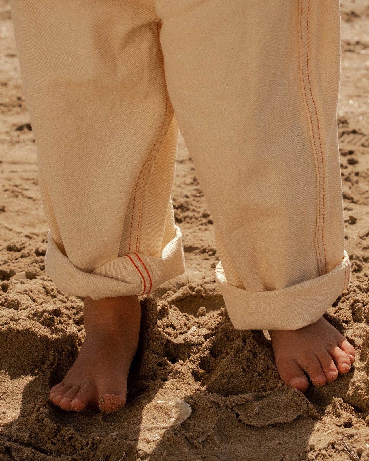 A close-up shows a child’s bare feet on sand, wearing GROWN - Organic Everyday Denim Pant Milk by GROWN with the rolled-up relaxed fit bottoms visible.
