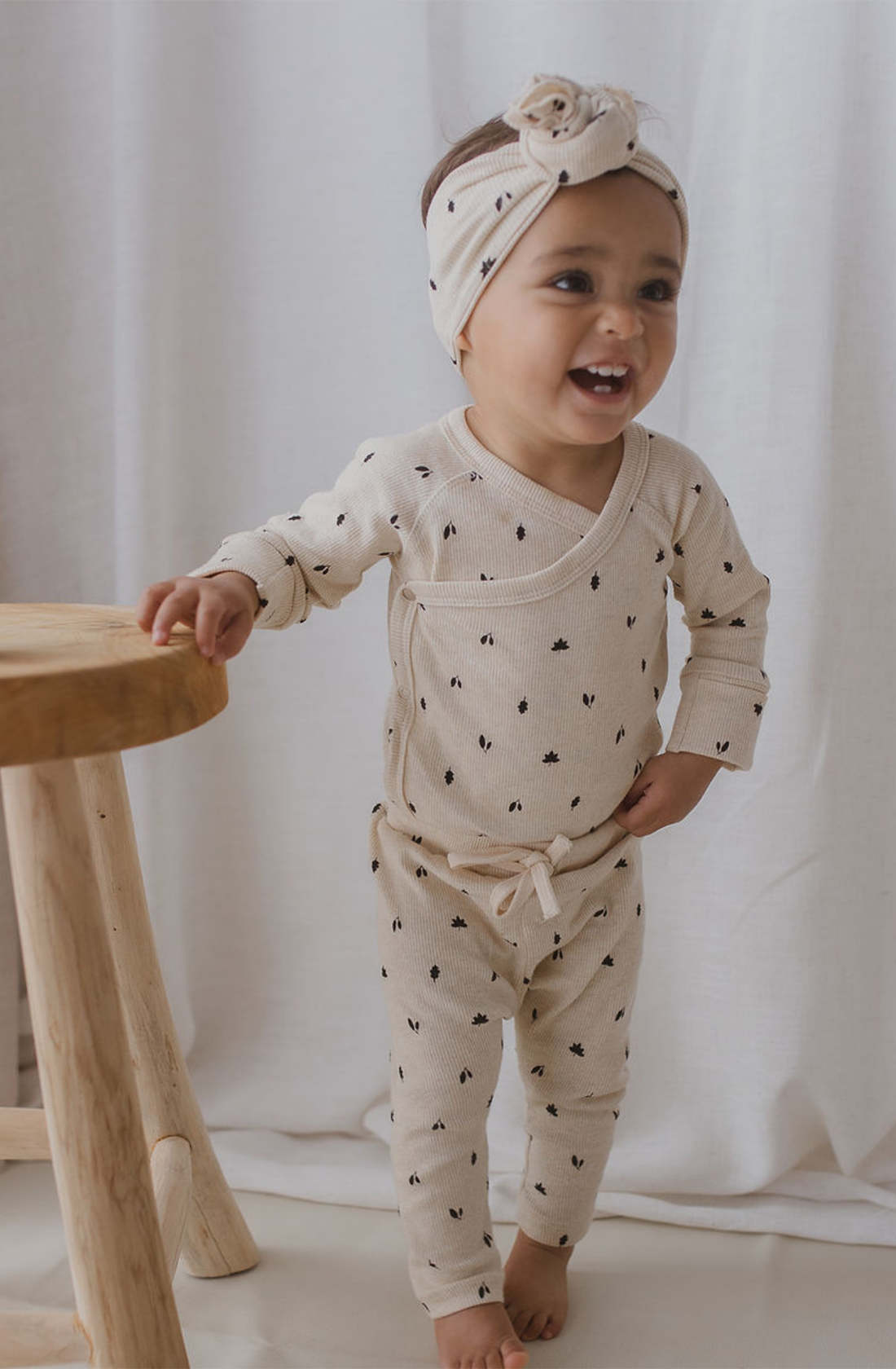 A toddler stands smiling while holding onto a wooden stool, dressed in a cream-colored organic cotton outfit with small black dot patterns. They wear a long-sleeve top, matching pants, and the SUSUKOSHI Elastic Headband Fall Leaves with an adorable bow. A white curtain serves as the background.