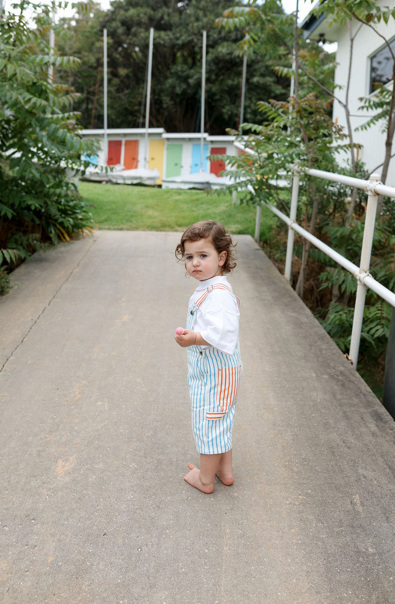 Toddler turned to the side wearing the Hola Ninch Overalls in blue stripe with red stripe straps and pocket details