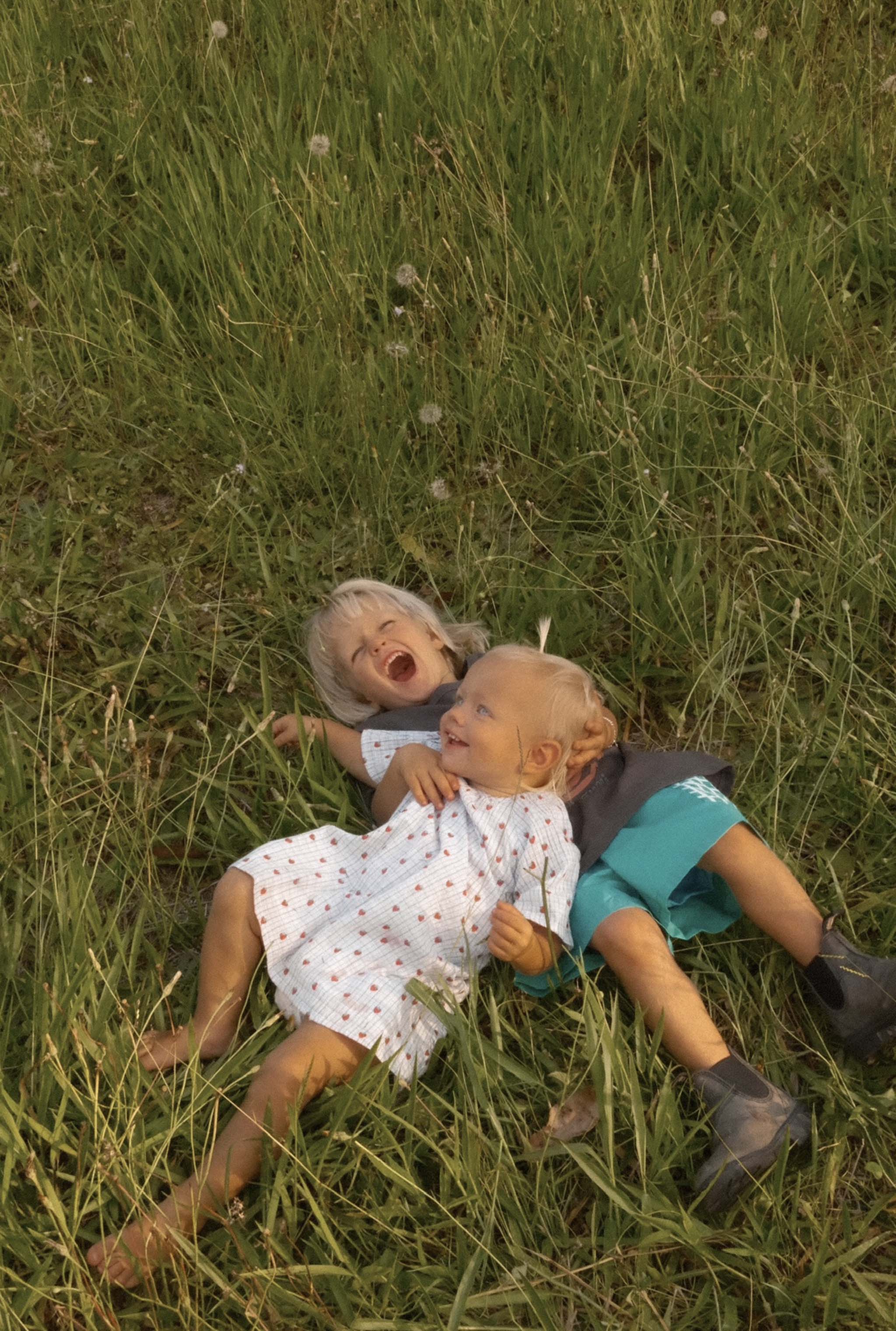 Two young children laugh joyfully on green grass, one in JUNI JNR's Penny Dress Strawberry Check - Size 4, the other in a gray shirt with blue shorts, surrounded by scattered dandelions as they relish their playful outdoor moment.