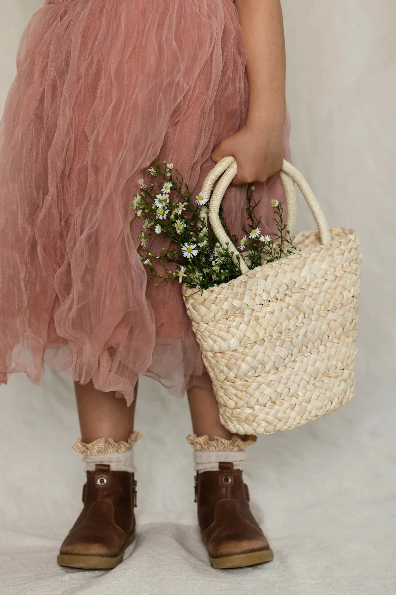 A child wearing the Bangalow Tutu Dress in Antique Rose by VALENCIA BYRON BAY and brown ankle boots with ruffled socks is holding a woven straw handbag filled with small white flowers. The image captures the child from the neck down, focusing on the outfit and accessories.