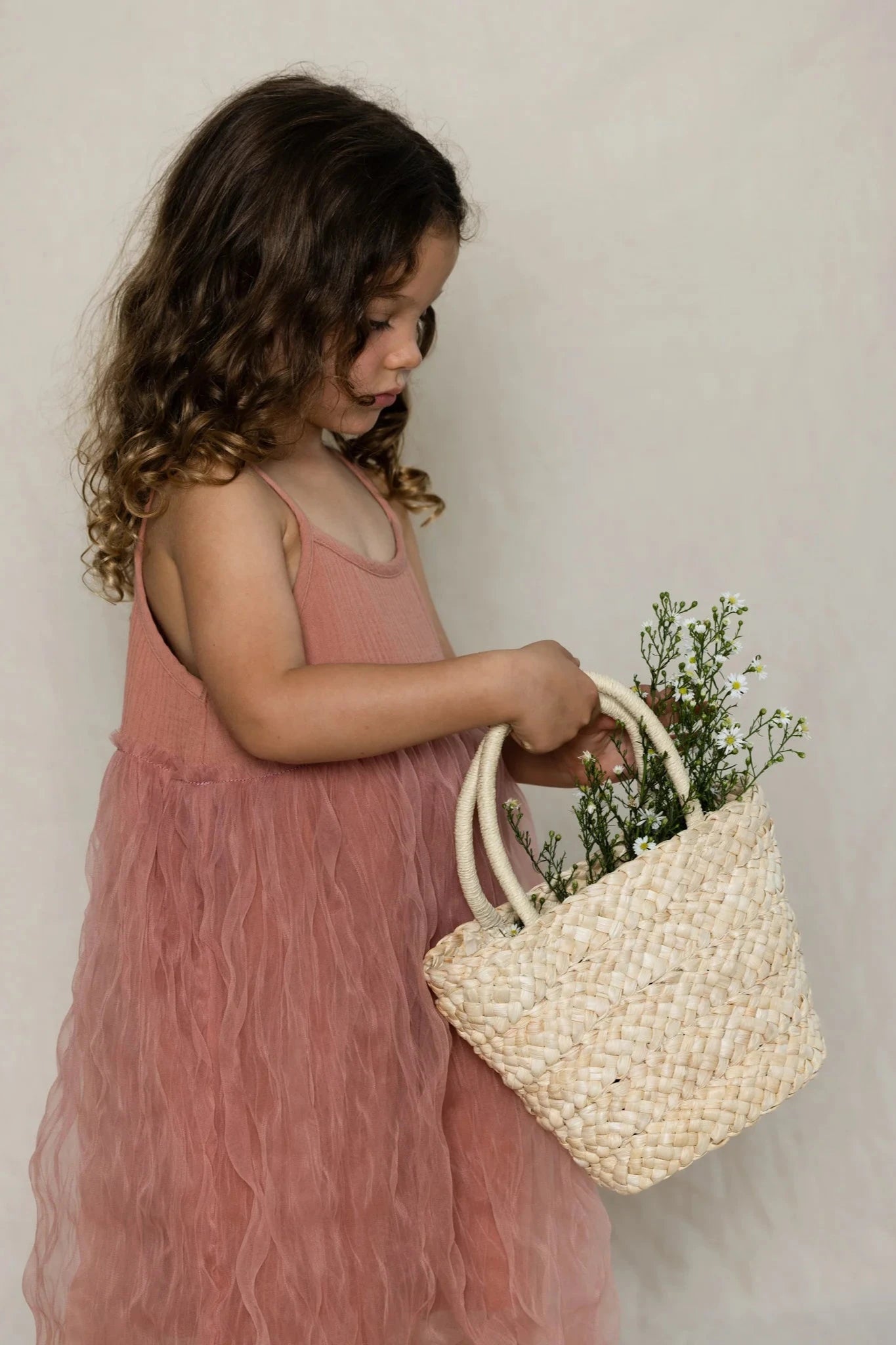 A young girl with curly hair, wearing a sleeveless VALENCIA BYRON BAY Bangalow Tutu Dress in Antique Rose, is holding a woven basket filled with greenery. She looks down at the basket while standing against a light, neutral background.