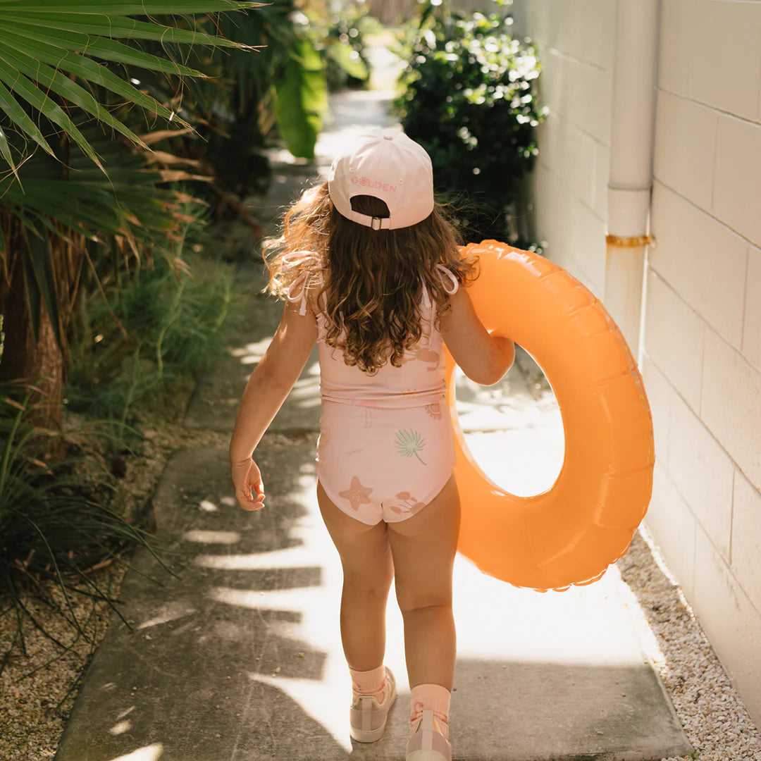 A young girl with curly brown hair walks along a path, dressed in a GOLDEN CHILDREN Palm Beach One Piece Swimsuit in Palm Beach Pink, crafted from recycled nylon, along with a coordinating cap. She carries an orange inflatable ring and is enveloped by green plants as sunlight filters through the leaves.