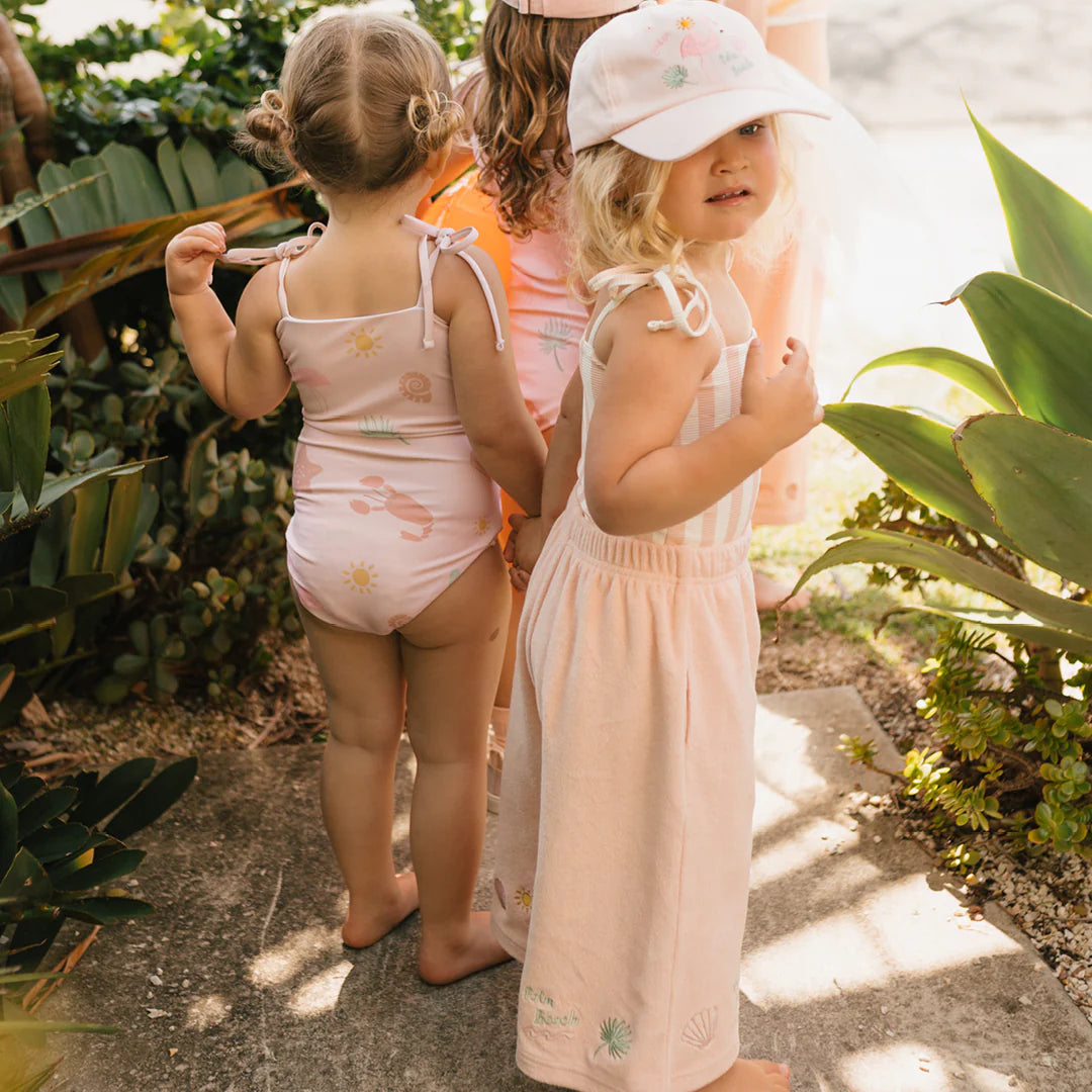 Three young children stand outdoors near lush green plants. The cheerful scene showcases one child in a Palm Beach One Piece Swimsuit Palm Beach Pink by GOLDEN CHILDREN with adjustable shoulder ties, another in a dress, and the last in a pink hat—all embracing the sunny day in light-colored summer clothing.