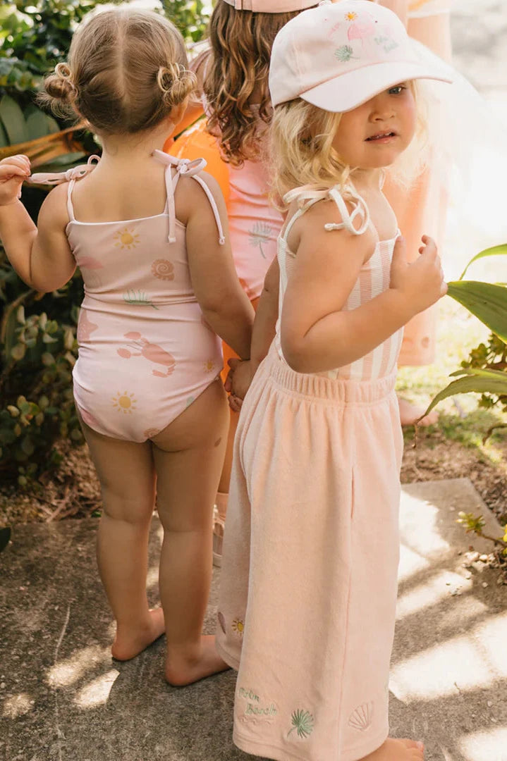 Two young children stand outdoors. One wears a pink swimsuit featuring sun and flower patterns, while the other sports a matching pink dress paired with the Palm Beach Cap in Palm Beach Pink by GOLDEN CHILDREN. Both have light-colored hair and smile in the sunny, green setting.