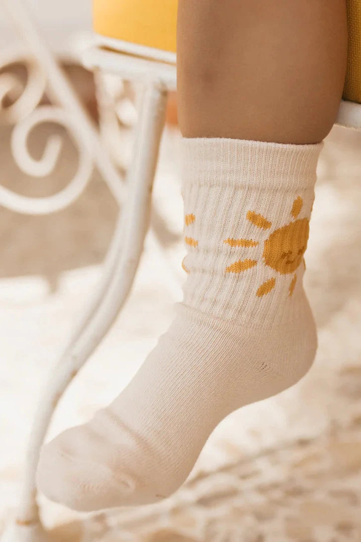 A child is wearing Golden Sun Socks in Tan, designed by GOLDEN CHILDREN, featuring a vibrant Golden Sun motif on the side. The child's foot rests comfortably on a white metal chair, while the softly blurred background in warm tones suggests a cozy setting.