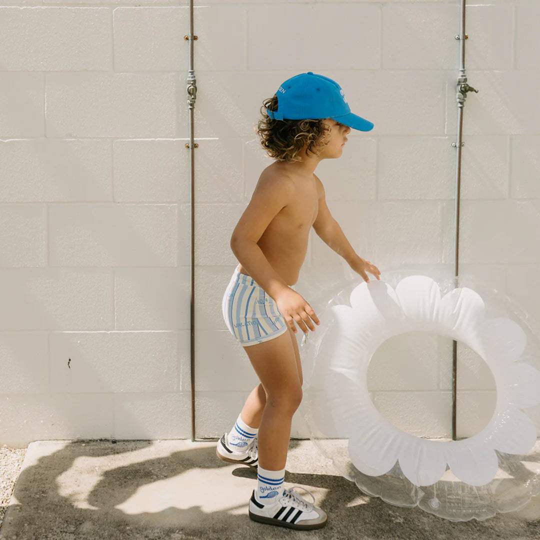 A young child sporting the GOLDEN CHILDREN Vacation Swim Trunks in a Boca Raton Stripe design, alongside a matching cap, socks, and sneakers, holds a clear flower-shaped inflatable pool toy. They stand against a light-colored brick wall, gearing up for exciting aquatic adventures.