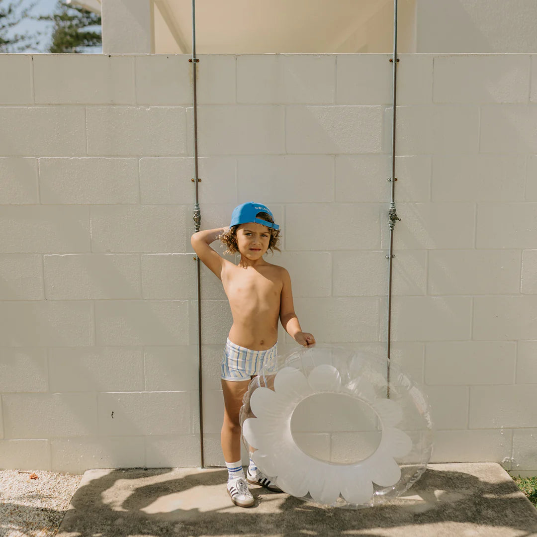 A young child wearing the "Vacation Swim Trunks Boca Raton Stripe" by GOLDEN CHILDREN, along with sneakers and a cap, stands against a white brick wall. The child holds a transparent, flower-shaped inflatable ring and playfully poses with one hand behind their head. The sunny scene captures the essence of carefree outdoor fun.