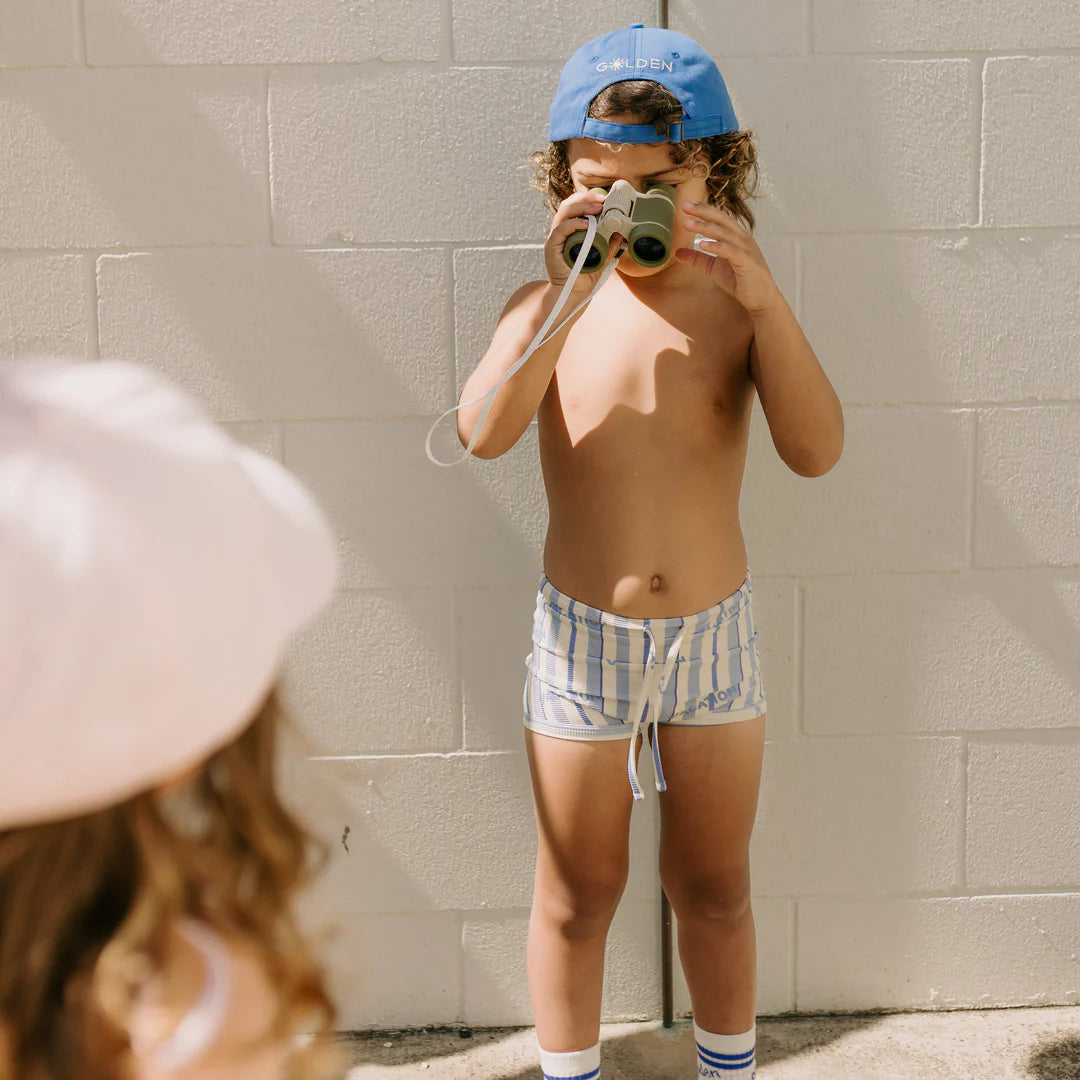 A child sporting the GOLDEN CHILDREN Vacation Swim Trunks in Boca Raton Stripe, along with a blue cap and socks, holds binoculars while standing against a white brick wall. In the foreground, another child is partially visible in a pink cap. The sunny and playful scene captures the essence of a fun vacation print.