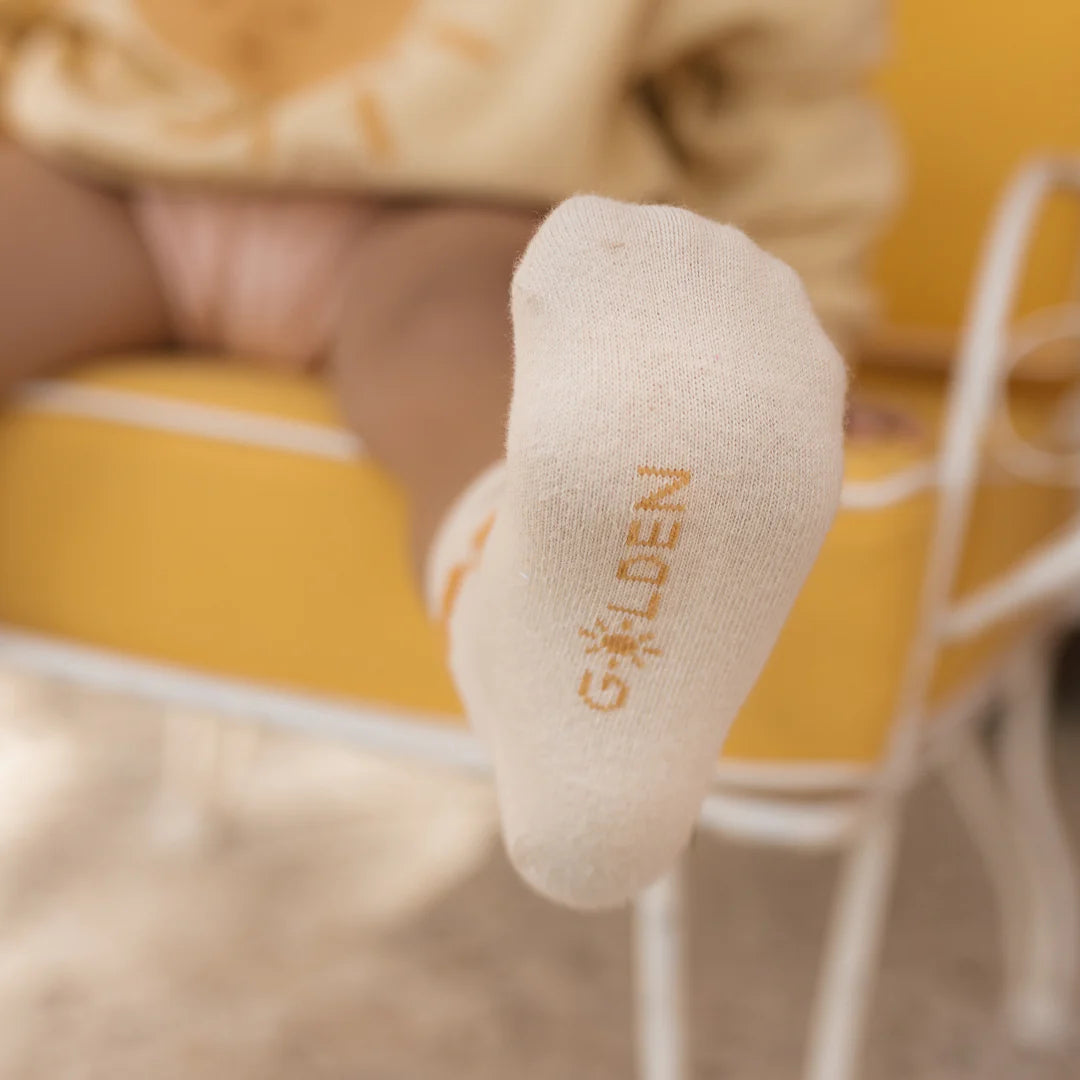 A close-up captures a toddler's foot adorned with GOLDEN CHILDREN's Golden Sun Socks in tan. The child sits comfortably on a yellow chair, while the softly blurred background enhances the cozy ambiance.