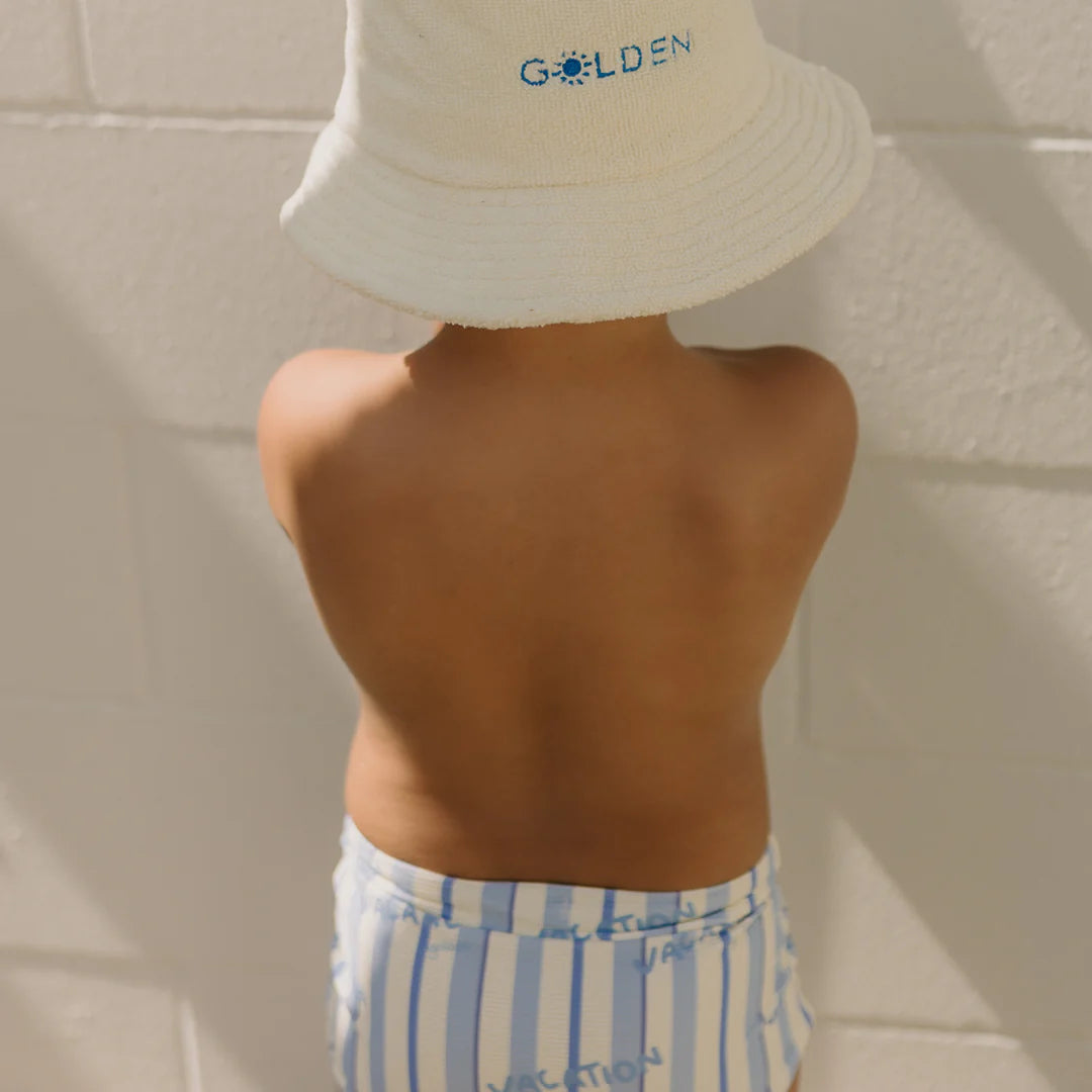 A child wearing a white bucket hat with the "GOLDEN CHILDREN" label stands facing a wall, their bare back exposed. They are dressed in Vacation Swim Trunks Boca Raton Stripe, crafted from breathable swimwear fabric. The striped design shines brightly in the sunlight, creating playful shadows on the wall.