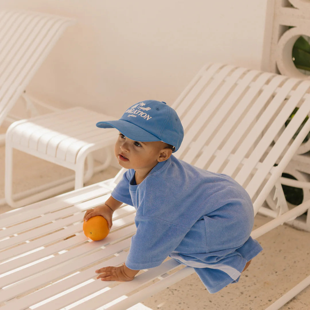 A toddler wearing a 100% cotton blue outfit and the stylish On Vacation Cap in Deep Sea Blue by GOLDEN CHILDREN is seated on a white chaise lounge. The child, holding an orange, gazes off to the side in the sunny outdoor setting. The cap features an adjustable brass clasp for perfect fit and comfort.