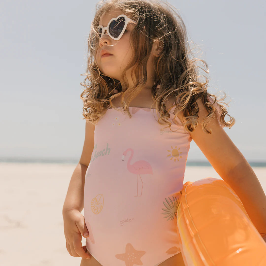 A young girl with curly hair stands on the beach wearing heart-shaped sunglasses and a Palm Beach One Piece Swimsuit Palm Beach Pink by GOLDEN CHILDREN, crafted from recycled nylon, featuring adjustable shoulder ties and a flamingo design. She holds an orange inflatable float as she gazes at the ocean under a clear blue sky.