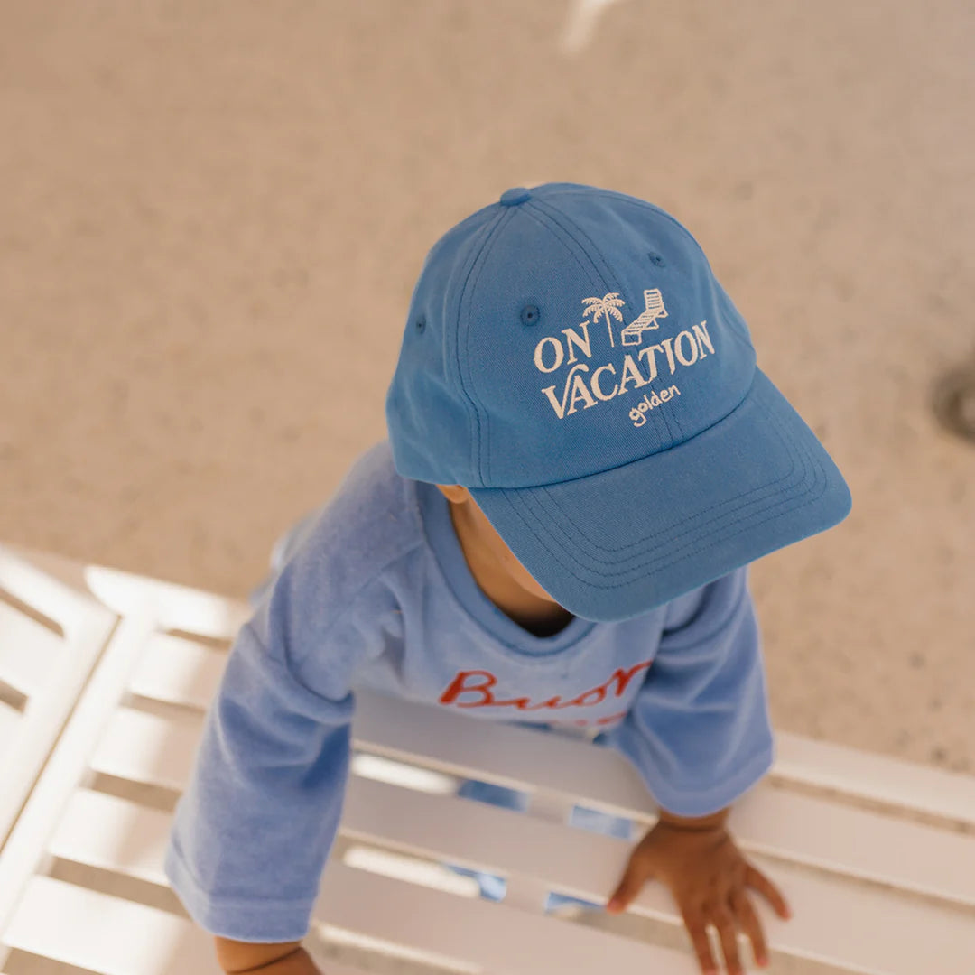 A child wearing the GOLDEN CHILDREN "On Vacation Cap" in Deep Sea Blue, complete with an adjustable brass clasp and made from 100% cotton, stands at a white slatted surface. This stylish accessory perfectly matches the child's blue shirt with some red text visible. The background features a light, neutral color.