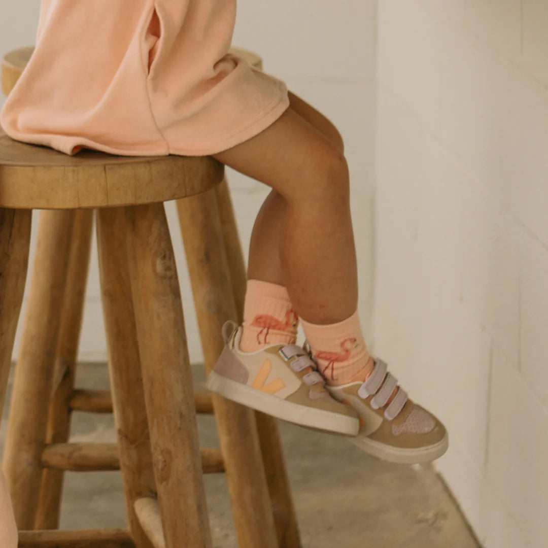 A child sits on a wooden stool, wearing a peach-colored dress paired with Flamingo Socks Sunset Pink from GOLDEN CHILDREN. Beige sneakers with reinforced toe caps and Velcro straps complete the look, set against a light-colored wall and concrete floor.