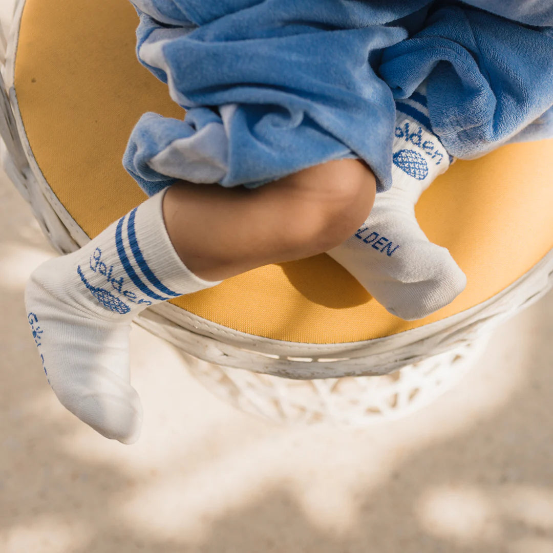 A baby wrapped in a soft blue blanket sits cross-legged on a yellow cushion, embodying the Palm Beach lifestyle. The little one sports GOLDEN CHILDREN's Retro Boy Socks Vanilla—white with blue stripes and a charming logo—adding a dash of style to the cozy scene.