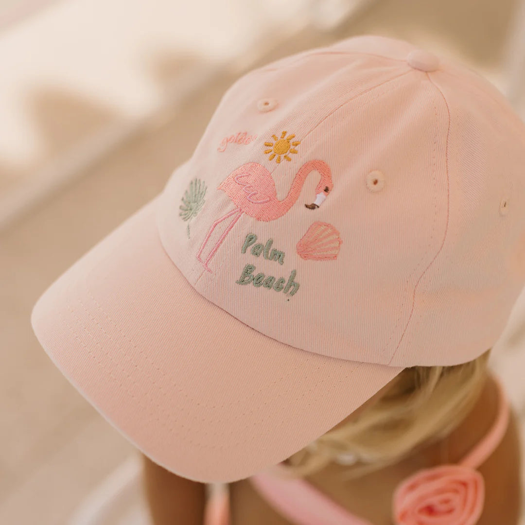 A person wearing the Palm Beach Cap in Palm Beach Pink by GOLDEN CHILDREN, made from 100% cotton, features an embroidered flamingo, sun, and seashells. The words "Palm Beach" add charm to this pastel-colored outfit.