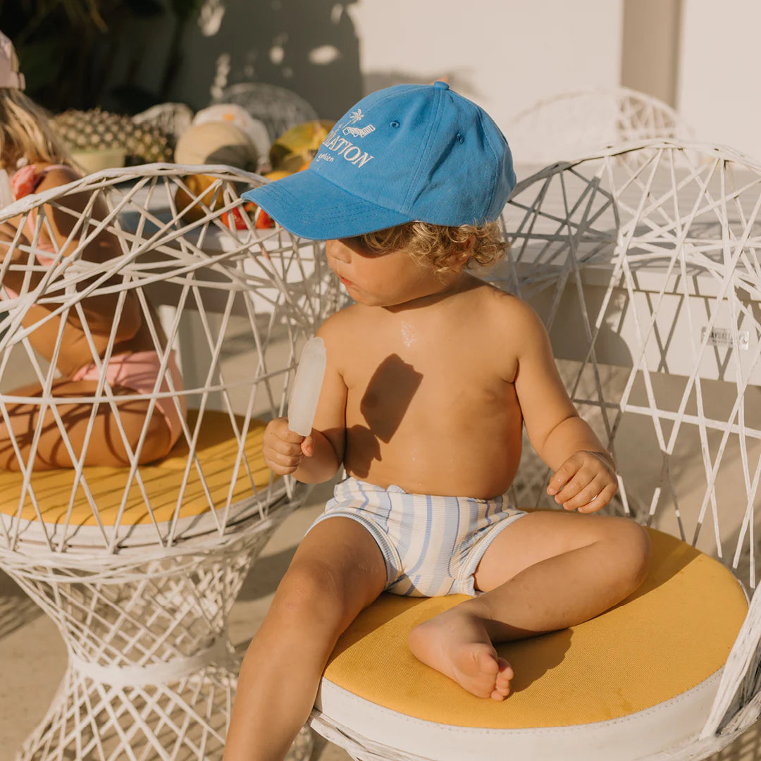 A toddler with curly hair sits on a white wire chair with yellow cushions, holding a popsicle. They are wearing GOLDEN CHILDREN's Vacation Swim Trunks Boca Raton Stripe and a blue cap, gazing to the side. Another child is partially visible in the background.