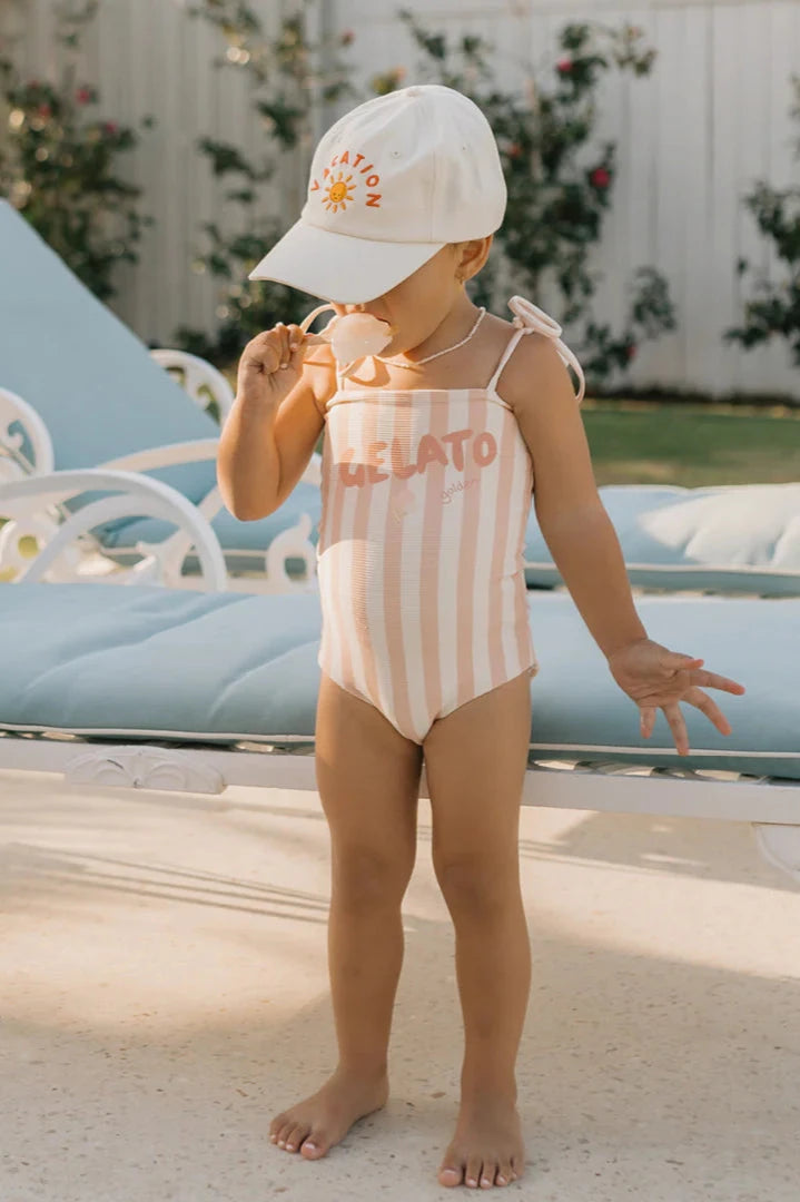 A child wearing the GOLDEN CHILDREN Gelato One Piece Swimsuit in Rose/Buttercream with adjustable shoulder ties and a sun hat that says "Vacation" stands by a poolside lounger, holding sunglasses. The playful gelato print adorns the ensemble, set against a background of a flower-adorned fence.