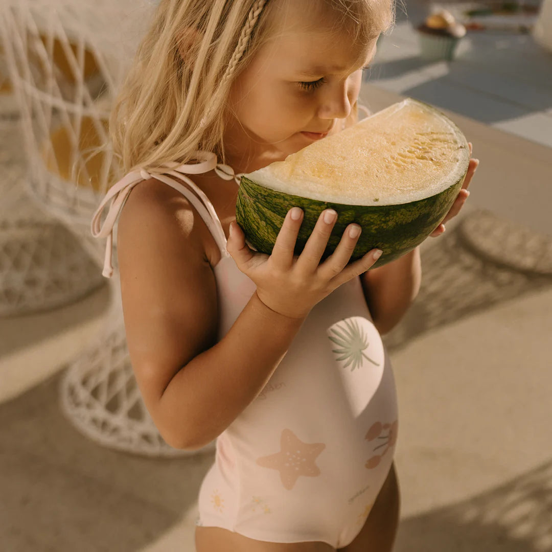 A young girl wearing a Palm Beach One Piece Swimsuit Palm Beach Pink from GOLDEN CHILDREN stands outdoors, adjusting the shoulder ties while enjoying the aroma of a large slice of watermelon. The scene is bright and sunny, with patio furniture visible in the background. Made from recycled nylon, her swimsuit perfectly complements the vibrant ambiance.