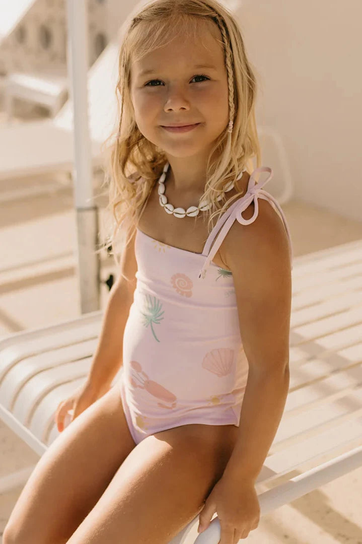 A young girl with blonde hair sits on a white lounge chair, wearing the Palm Beach One Piece Swimsuit in Palm Beach Pink by GOLDEN CHILDREN. This light pink swimsuit, made from recycled nylon, showcases beach-themed patterns and adjustable shoulder ties. Her look is complemented by a shell necklace against a sunny and bright background.