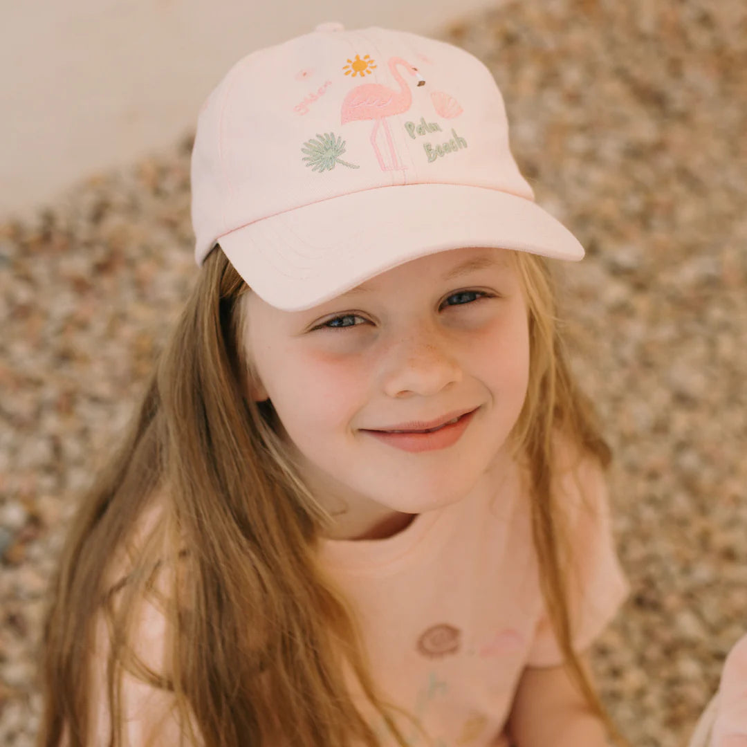 A smiling child wearing a 100% cotton light pink T-shirt and a Palm Beach Cap Palm Beach Pink from GOLDEN CHILDREN sits on gravel. The embroidered dad cap showcases a flamingo with the text "Palm Beach." The child has long hair and a cheerful expression.