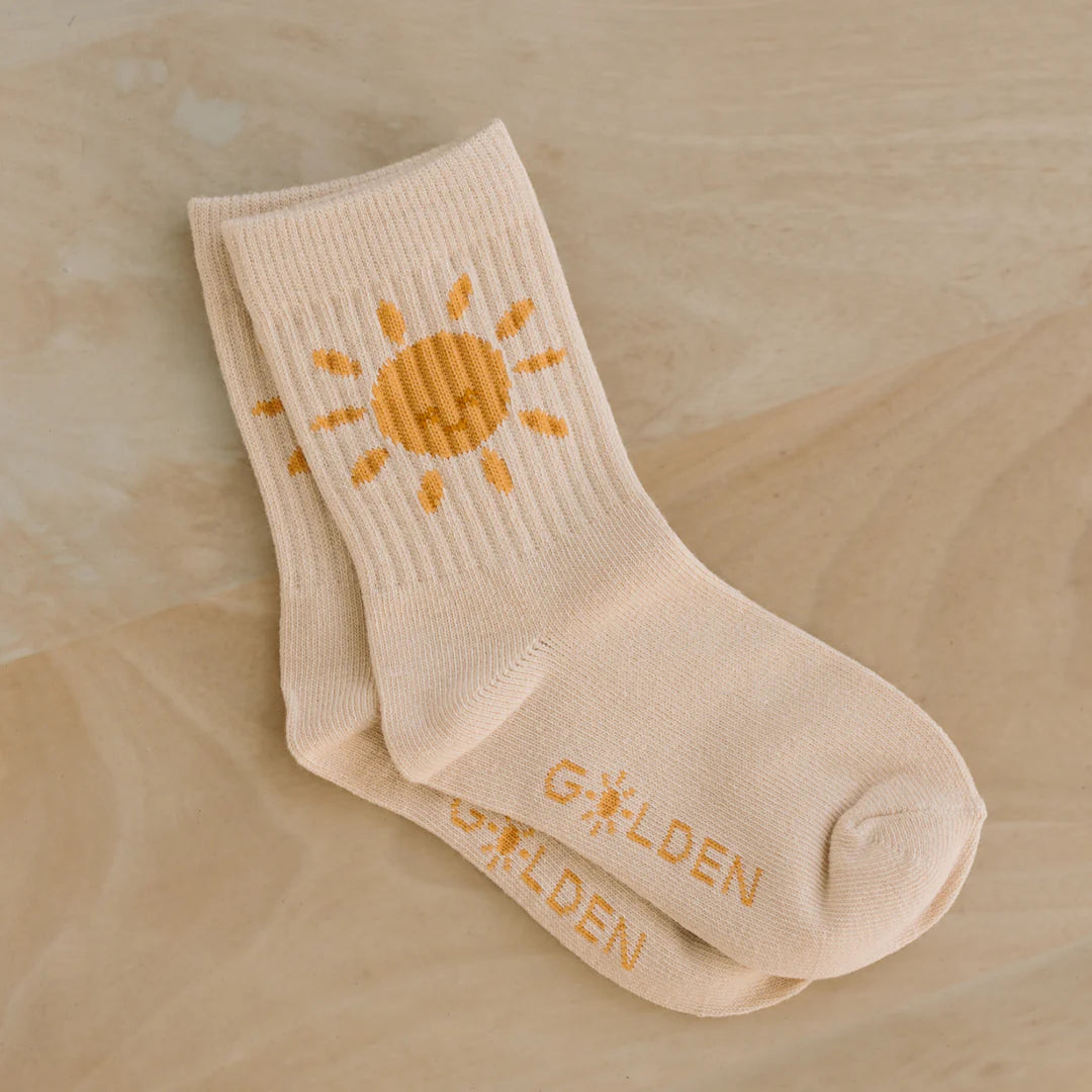 A pair of "Golden Sun Socks Tan" by GOLDEN CHILDREN, featuring a yellow sun design on the ankle and the word "GOLDEN" written in yellow near the reinforced toe caps, displayed on a light wood surface.