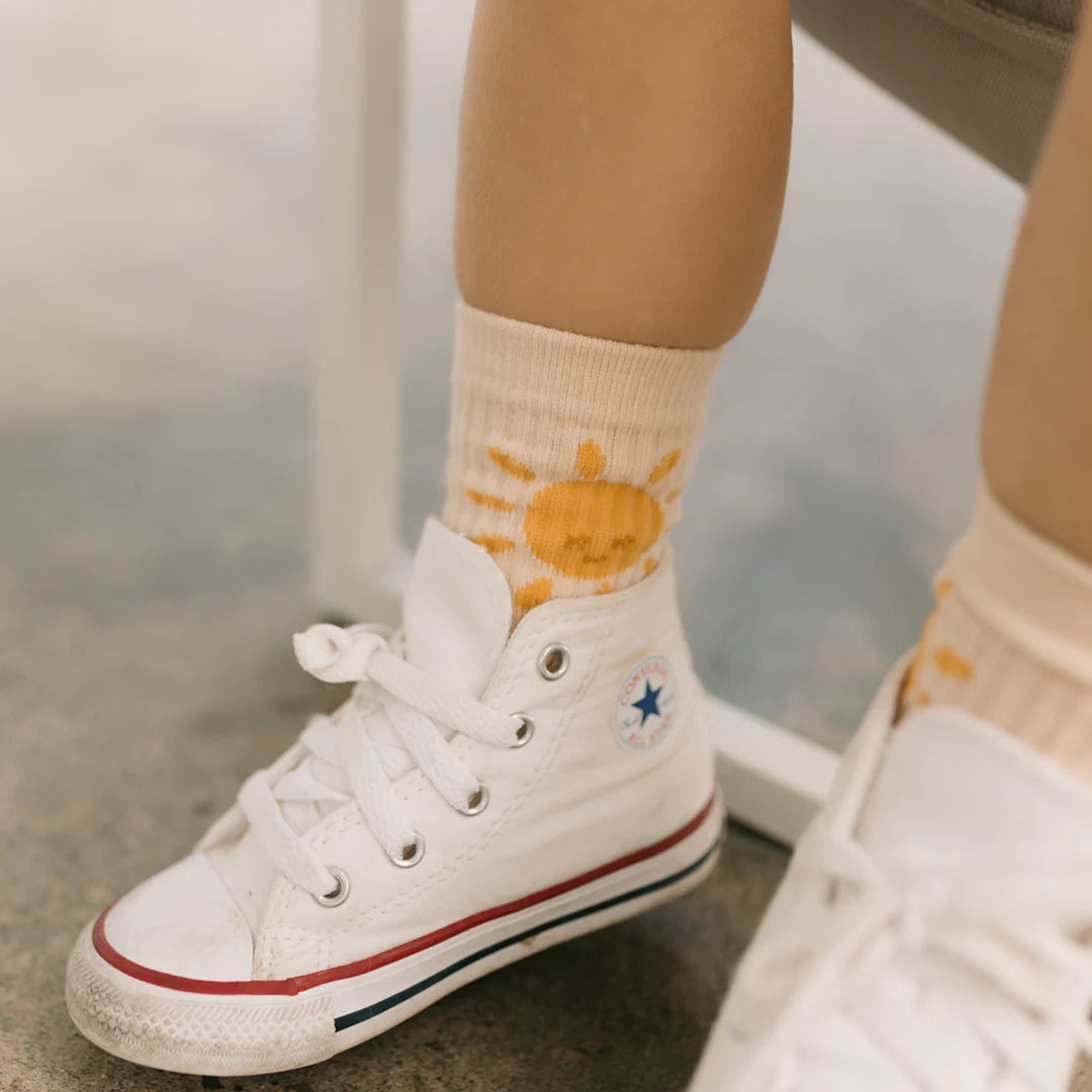 A close-up shows a child's leg adorned with white high-top sneakers featuring red and blue stripes and reinforced toe caps. The child sports Golden Sun Socks Tan by GOLDEN CHILDREN, vibrant in yellow with a sun design, against a blurred gray background.