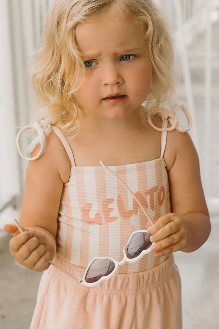 A young child with wavy blonde hair, wearing the GOLDEN CHILDREN Gelato One Piece Swimsuit in Rose/Buttercream, holds white sunglasses. The softly blurred background hints at an outdoor scene, capturing the essence of summer fun.