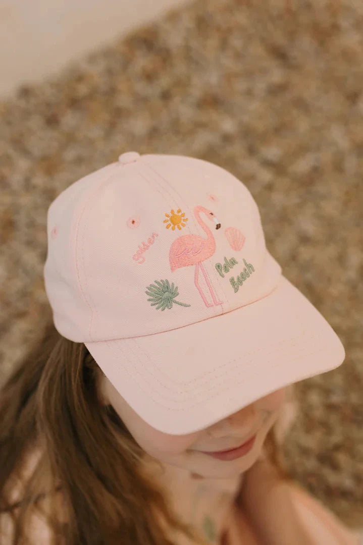 A young girl dons a Palm Beach Cap Palm Beach Pink from GOLDEN CHILDREN, crafted from 100% cotton and featuring embroidered designs of a flamingo and palm tree. The background of blurred pebbles adds to the cap's charm.