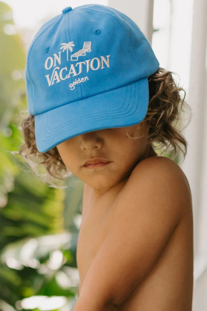 A child wearing the "On Vacation Cap Deep Sea Blue" from GOLDEN CHILDREN, with "ON VACATION" proudly stitched on it, looks down. Made from 100% cotton and featuring an adjustable brass clasp, the hat perfectly complements the sunlit green foliage backdrop, evoking a tropical vibe.