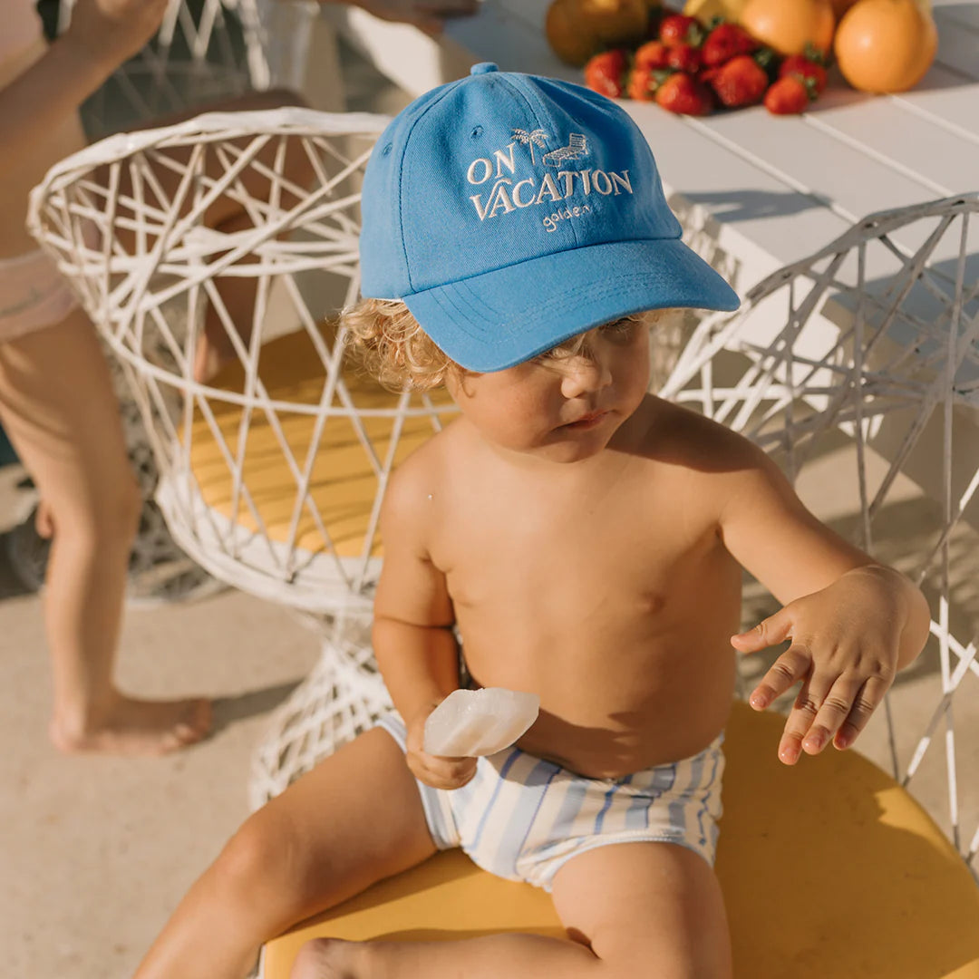 A toddler wearing a GOLDEN CHILDREN's On Vacation Cap in Deep Sea Blue, along with striped swim trunks, sits on a yellow bench. They are holding a piece of fruit and enjoying the sunny day. In the background, a table is filled with assorted fruits and another child is having fun. The cap comes equipped with an adjustable brass clasp for extra comfort.