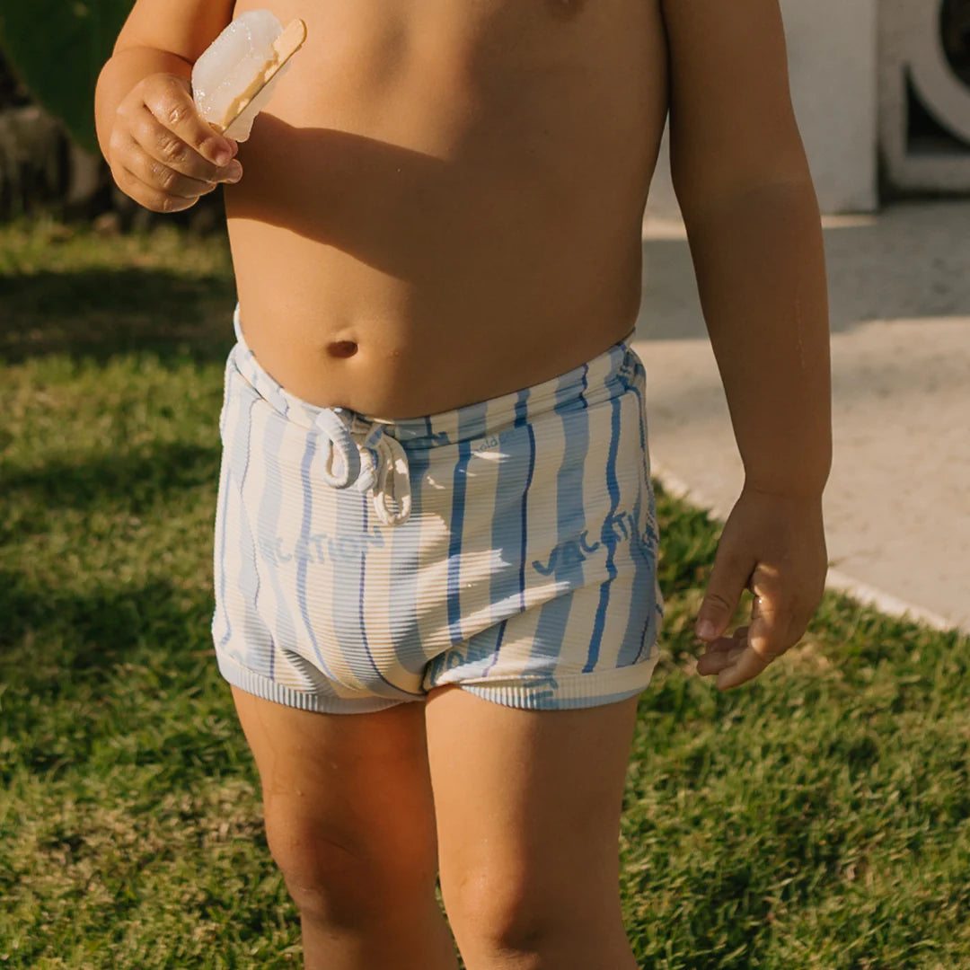 A child wearing the Vacation Swim Trunks Boca Raton Stripe from GOLDEN CHILDREN, adorned in a blue and white striped design made of stretchy swimwear fabric, holds a partially eaten ice cream bar. They stand on the grass, with sunlight casting shadows around them, and part of a building visible in the background.