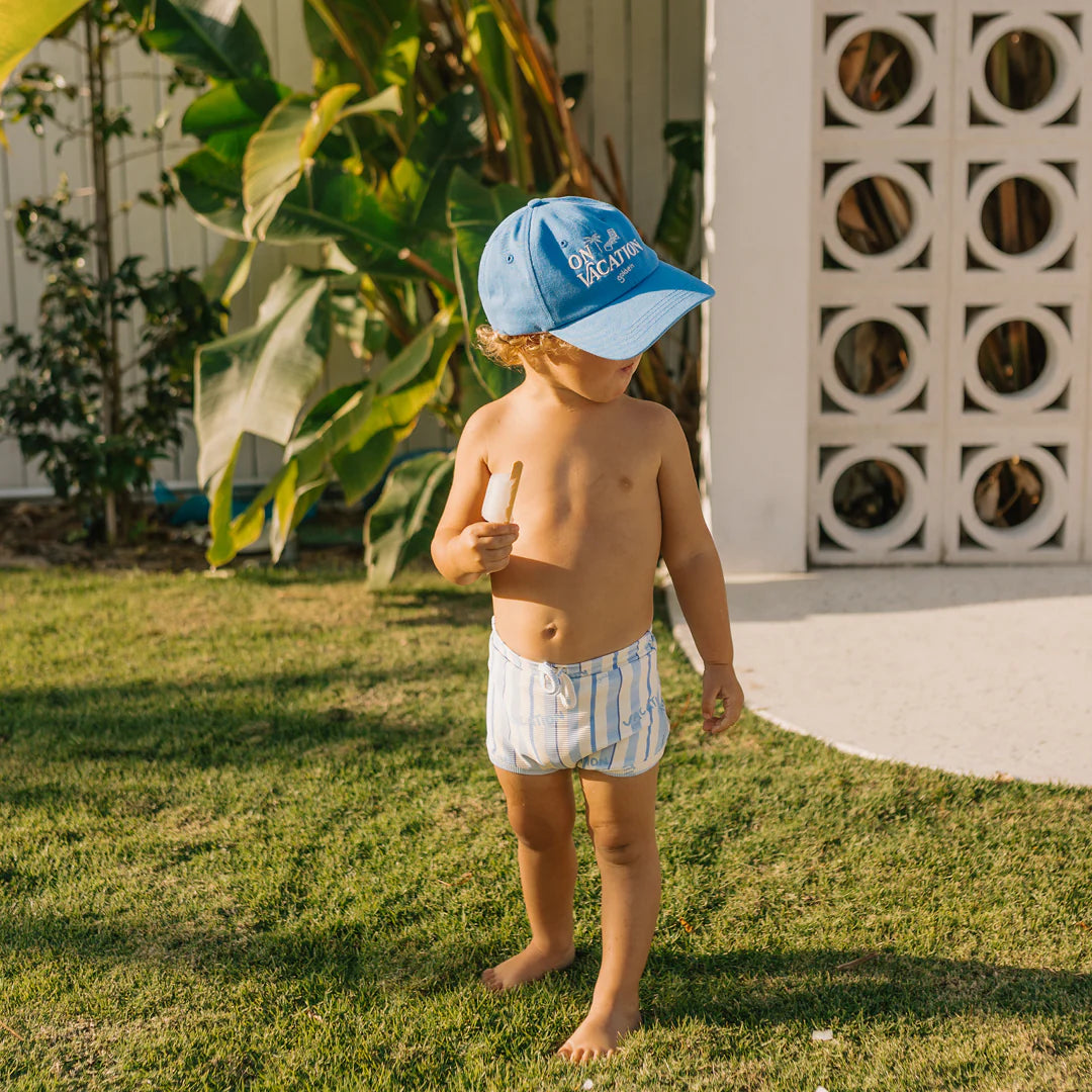 A toddler wearing the GOLDEN CHILDREN On Vacation Cap in Deep Sea Blue, featuring an adjustable brass clasp, pairs it with striped shorts while standing on grass and holding a stick. Sunlight creates shadows on their body, while lush green foliage and a white building with decorative patterns enhance the background.