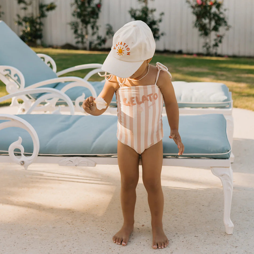 A toddler stands near a poolside lounge chair, wearing the Gelato One Piece Swimsuit in Rose/Buttercream by GOLDEN CHILDREN, featuring adjustable shoulder ties. The ensemble is complemented by a white cap adorned with a sun design. The child clutches a small snack in one hand, perfectly capturing the essence of a sunny day outdoors.