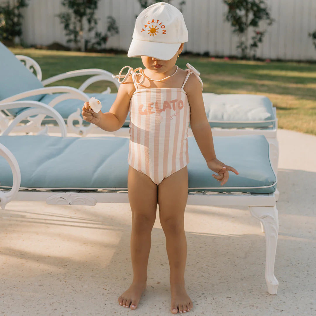 A toddler wearing the GOLDEN CHILDREN Gelato One Piece Swimsuit in Rose/Buttercream with adjustable shoulder ties and a cap featuring a sun design stands by a poolside lounge chair, clutching a seashell. A white fence and lush greenery form the background.