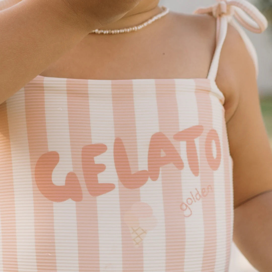 A child in the GOLDEN CHILDREN Gelato One Piece Swimsuit Rose/Buttercream, crafted from stretchy swimwear fabric and adorned with bold lettering, perfectly embodies a playful, summery vibe. The close-up features a necklace alongside an arm decorated with adjustable shoulder ties, enhancing the charm of its vibrant pink and white stripes.