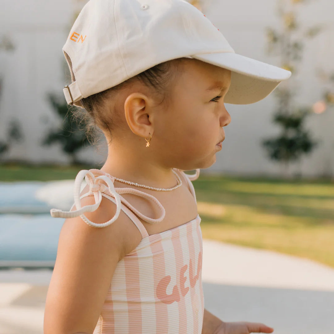 A toddler stands outdoors wearing the Gelato One Piece Swimsuit in Rose/Buttercream by GOLDEN CHILDREN, which features adjustable shoulder ties. The child looks to the side while sporting a beige cap adorned with tree patterns, set against a backdrop of blurred outdoor seating. The swimwear's stretchy fabric guarantees both comfort and style.