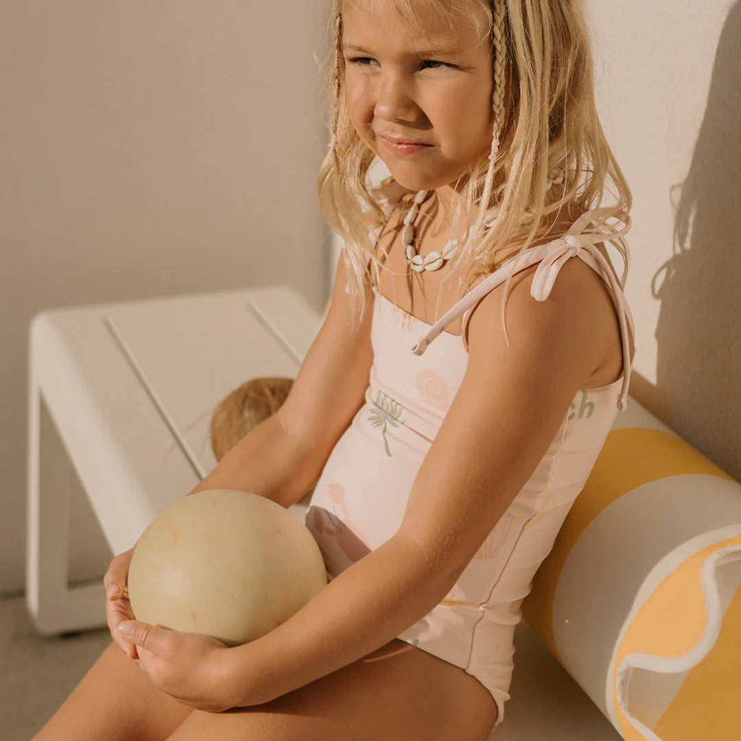 A young girl with wavy blonde hair sits on a white bench, wearing the light pink Palm Beach One Piece Swimsuit from GOLDEN CHILDREN, crafted from recycled nylon. She holds a small melon in her lap, and a striped cushion is beside her. Sunlight casts shadows on the background wall, drawing attention to the adjustable shoulder ties of her swimsuit.