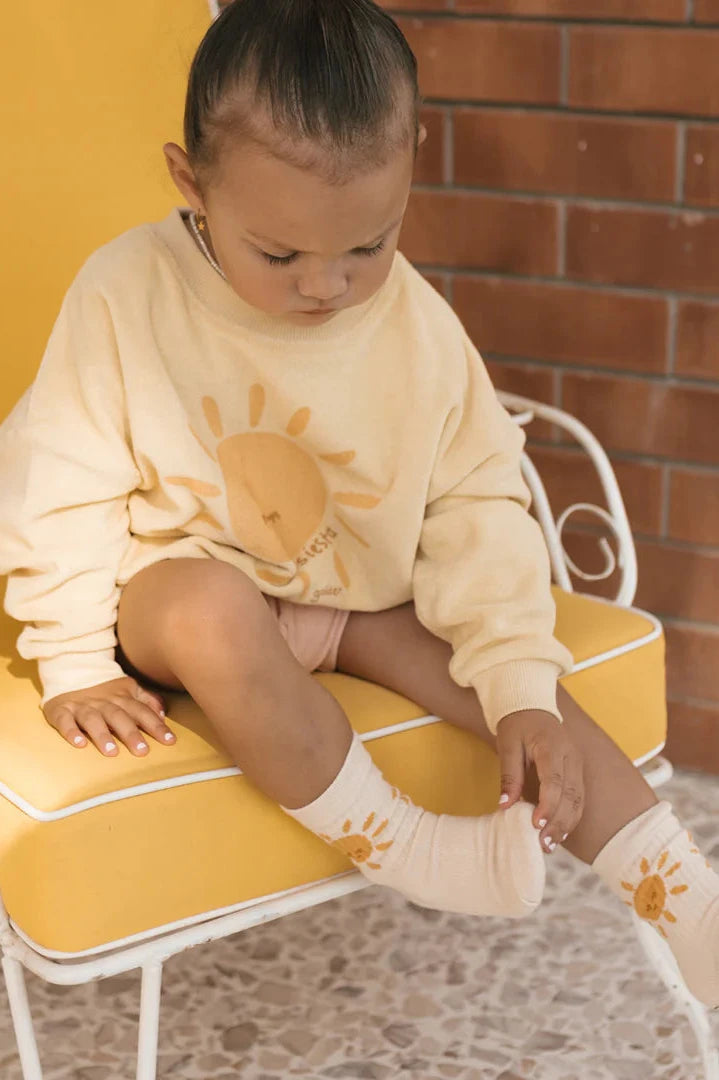 A toddler wearing a cream sweater with a Golden Sun design and coordinating Golden Sun Socks Tan by GOLDEN CHILDREN is seated on a yellow chair. They are looking down and adjusting their sock, against a backdrop of brick wall.