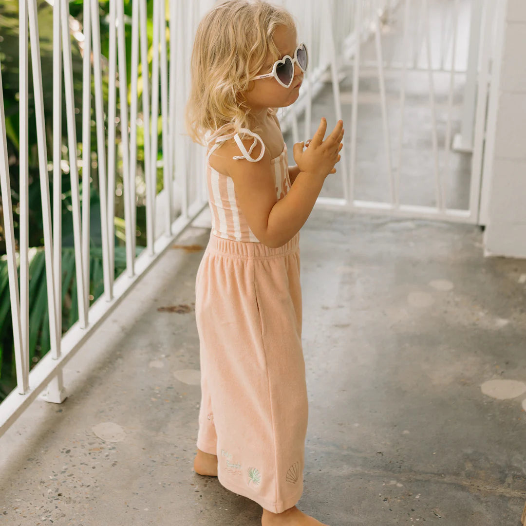 A young child with blonde hair stands on a balcony, exuding charm in heart-shaped sunglasses and a Gelato One Piece Swimsuit Rose/Buttercream by GOLDEN CHILDREN. The child is barefoot, basking in the sunny day that echoes the lively vibe of a vibrant gelato print.