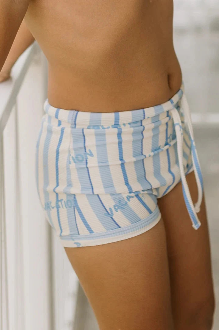 A person stands beside a white railing, modeling the Vacation Swim Trunks Boca Raton Stripe by GOLDEN CHILDREN. These stylish shorts flaunt playful blue and white stripes and have a subtle "LOVE" design intricately woven into the fabric.