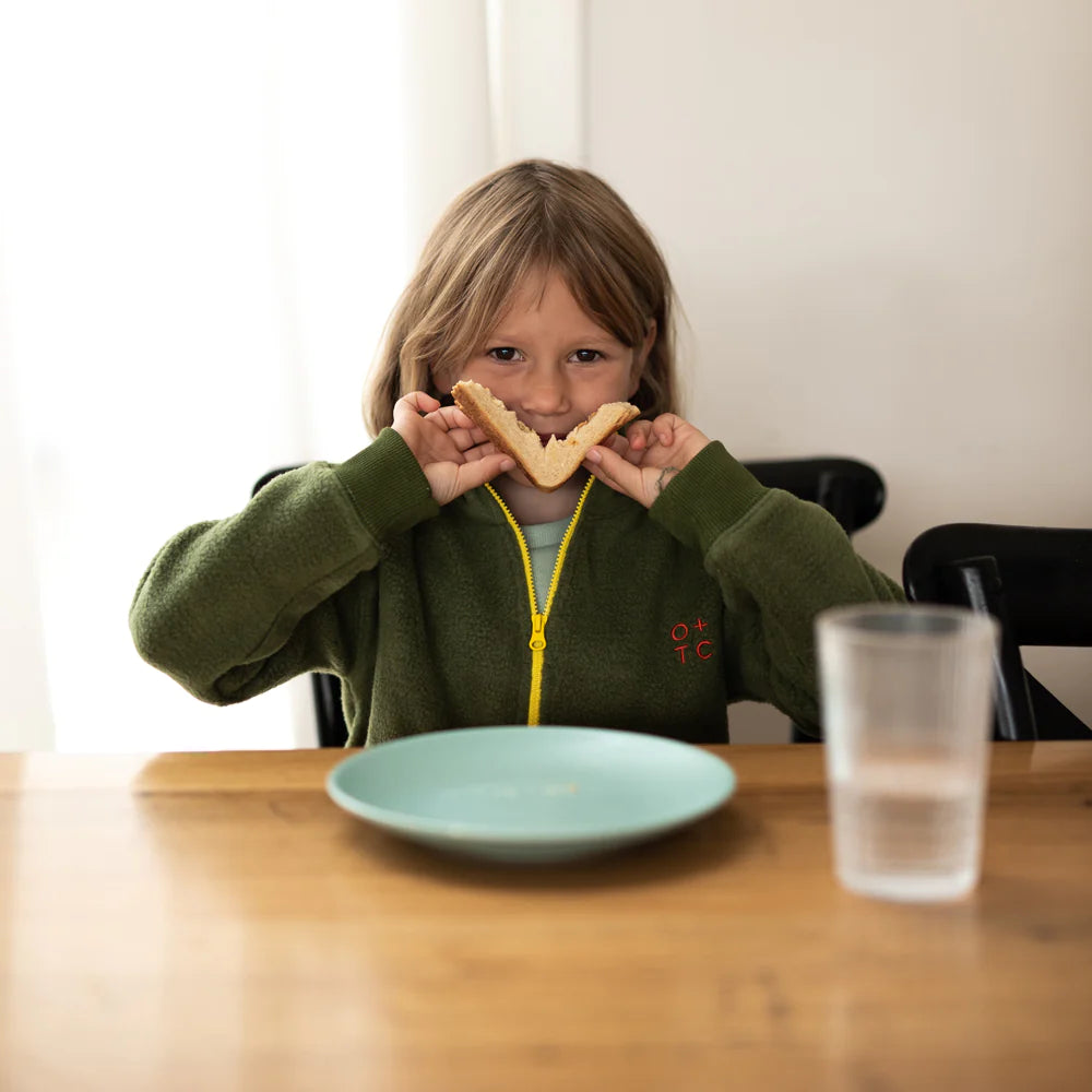 A young child with long hair and an Olive Zip Pullover, made from premium cotton by OLIVE + THE CAPTAIN, sits at a wooden table holding two triangular sandwich halves up to their face, forming a smiley face. In front of them, there's an empty light blue plate and a clear plastic cup. The background is plain and neutral.