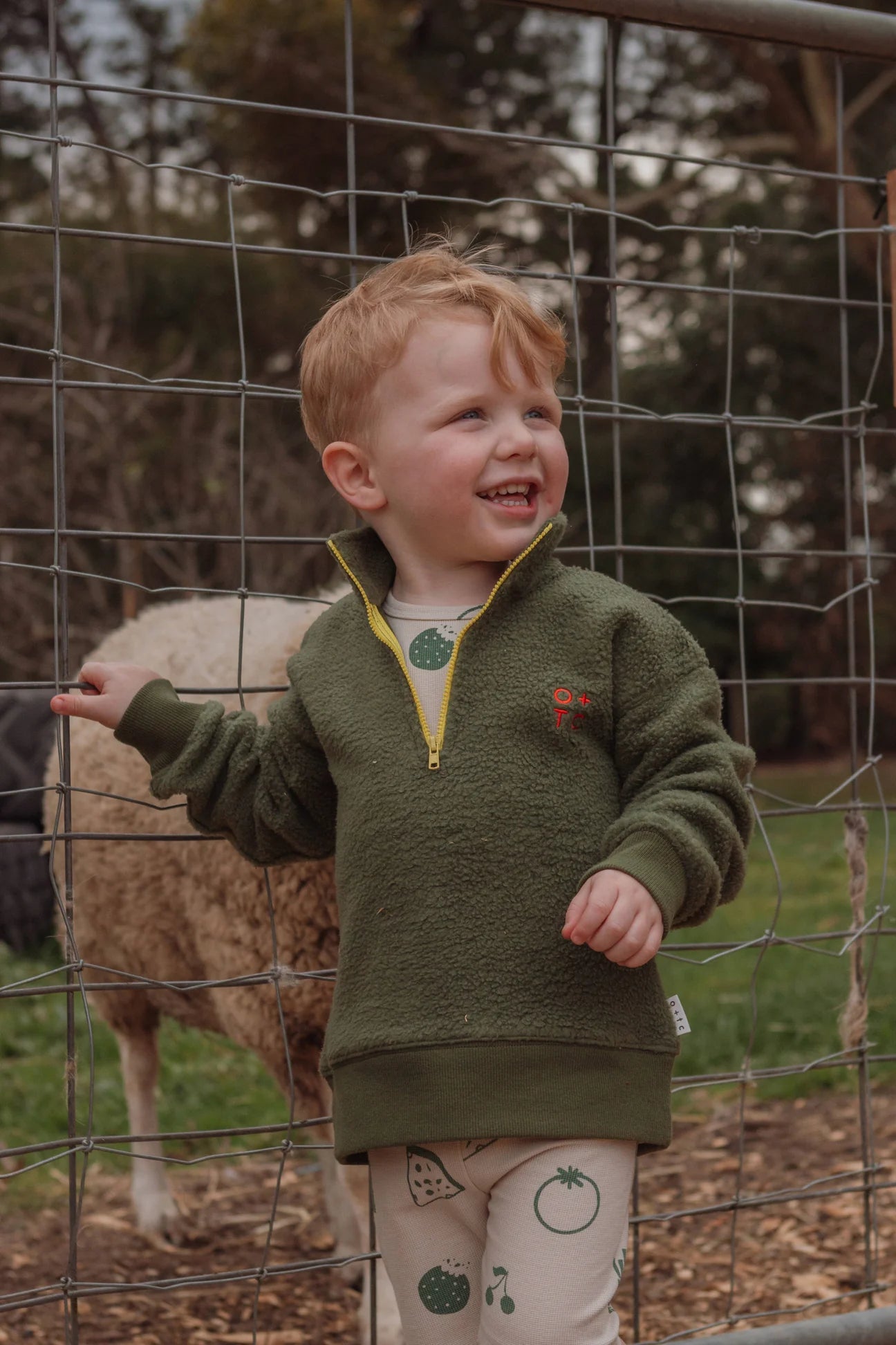 A young child with short, light-colored hair stands smiling while holding onto a wire fence. The child is wearing the Olive Zip Pullover by OLIVE + THE CAPTAIN and cream-colored pants adorned with vegetable prints, crafted from premium cotton. Behind the child, a sheep grazes in a grassy area surrounded by trees.