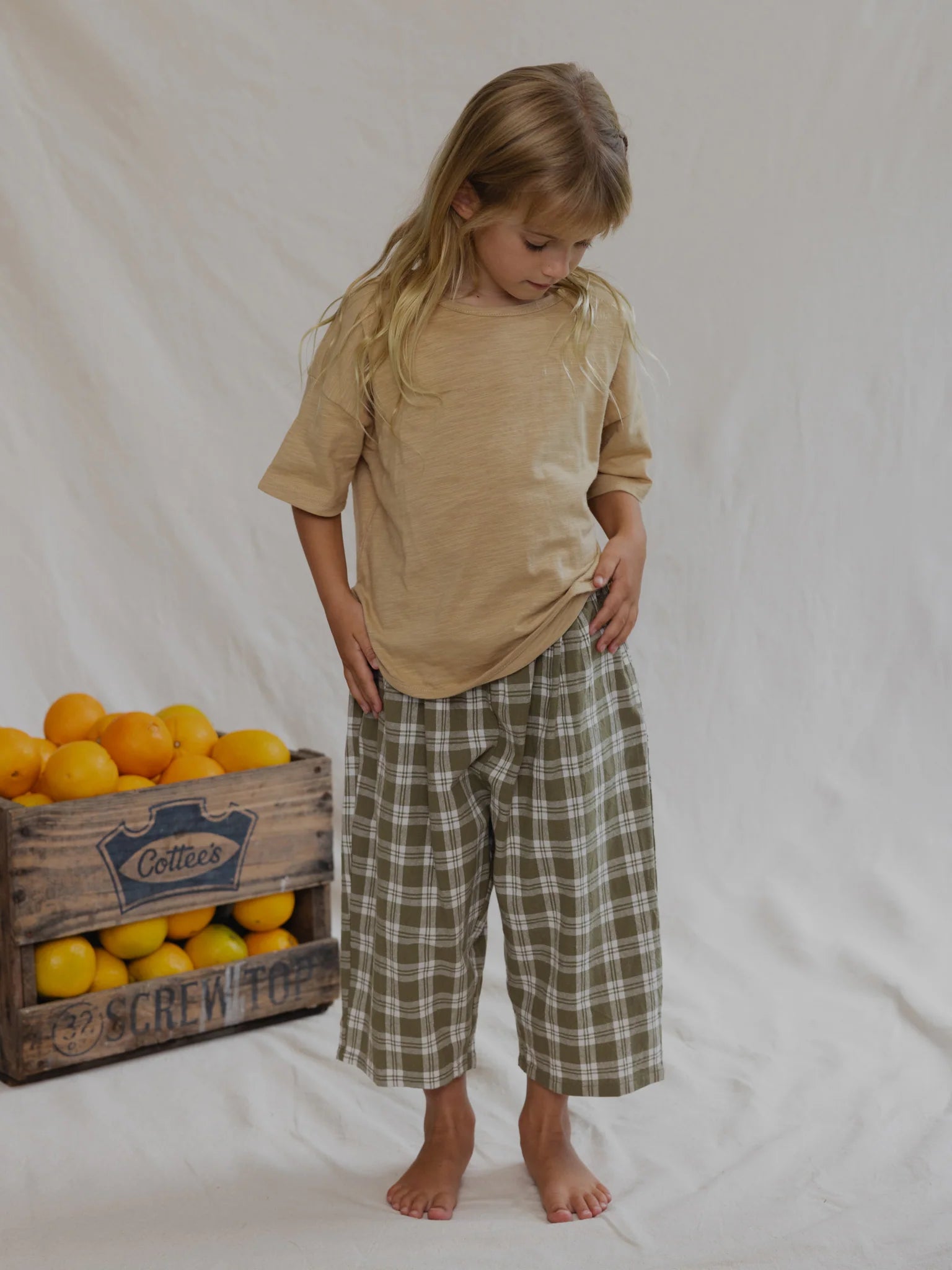 A young child with long blonde hair stands barefoot against a white cloth background. They are wearing a beige t-shirt and the Rio Pant Olive, 100% cotton and of unisex design by VALENCIA BYRON BAY. On the left side sits a wooden crate filled with oranges labeled "Cottes." The child is looking down at their pants.