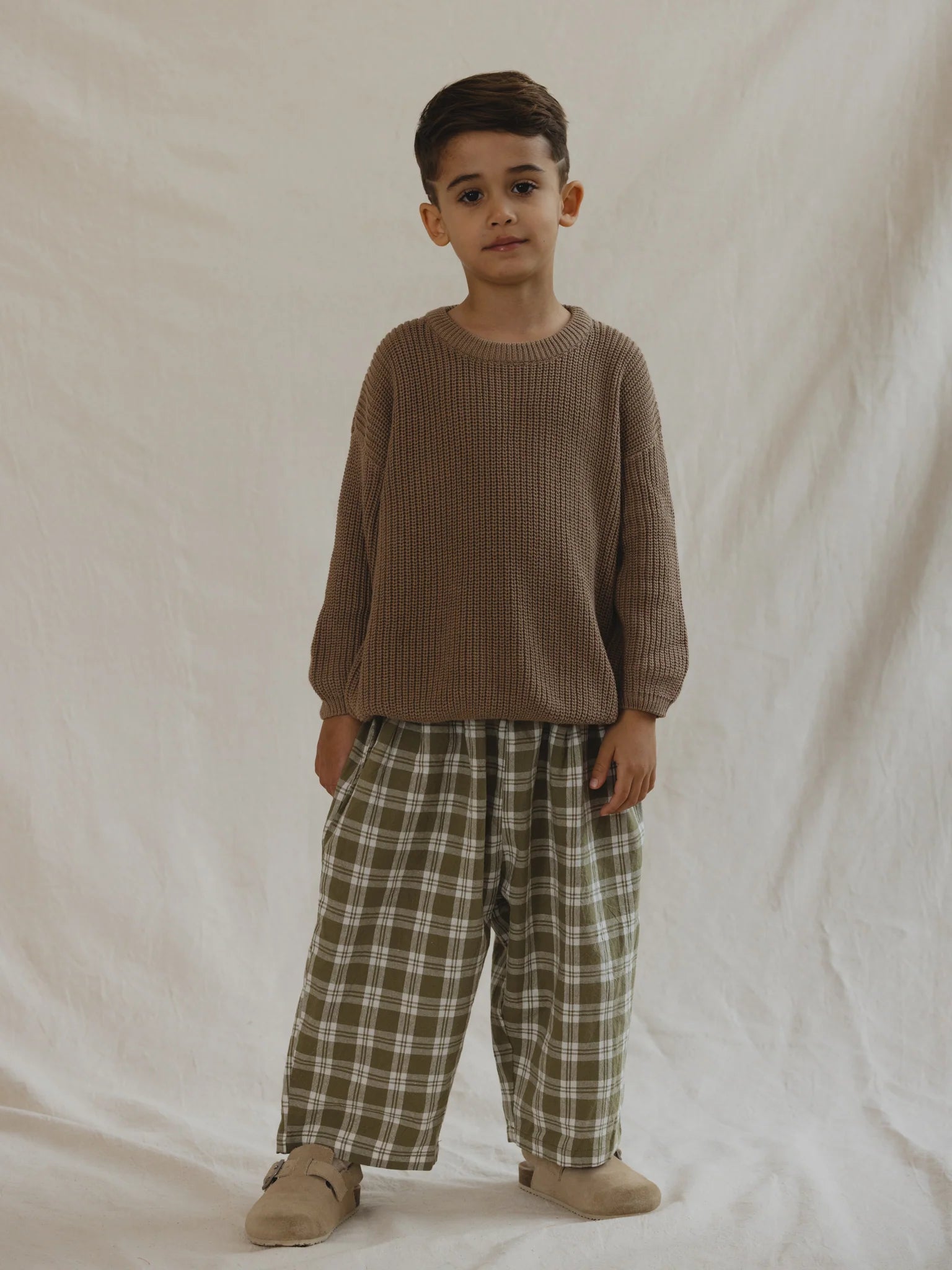 A young child stands against a plain, light-colored background. The child is wearing a loose-fitting, light brown sweater, VALENCIA BYRON BAY's Rio Pant Olive featuring an olive green print, and beige slipper-style shoes. They have short dark hair and a neutral expression.