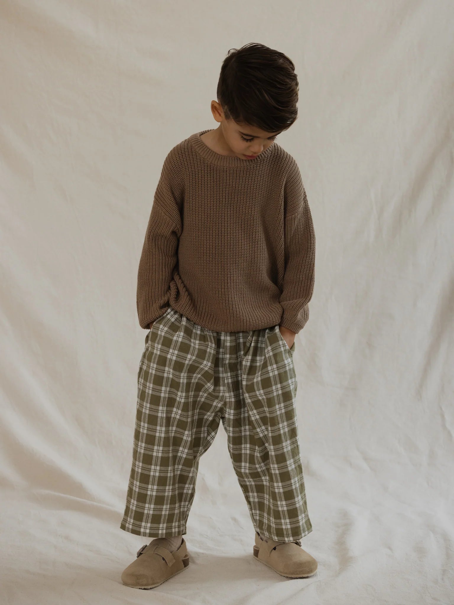 A young child stands with their hands in the pockets of their VALENCIA BYRON BAY Rio Pant Olive, wearing a loose-fitting, brown sweater and tan shoes. The 100% cotton plaid pants feature a unisex design with an olive green print that adds a subtle touch of style. The child looks downwards and is set against a neutral, light-colored background.