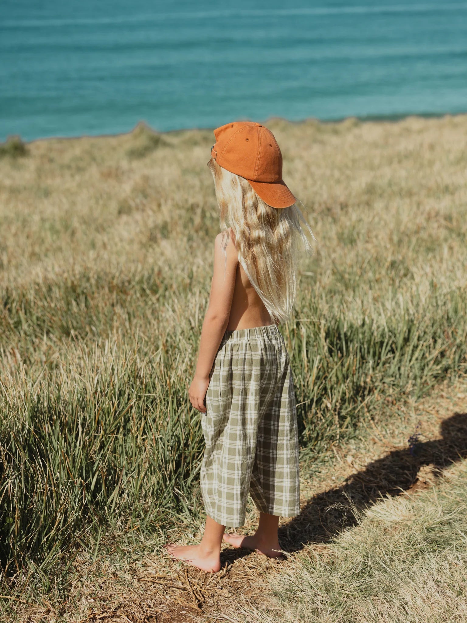 A child with long blonde hair, wearing a burnt orange cap and the VALENCIA BYRON BAY Rio Pant in olive, stands barefoot on a grassy area facing the sea. The background is a serene view of the turquoise ocean and an expansive field under a clear sky.