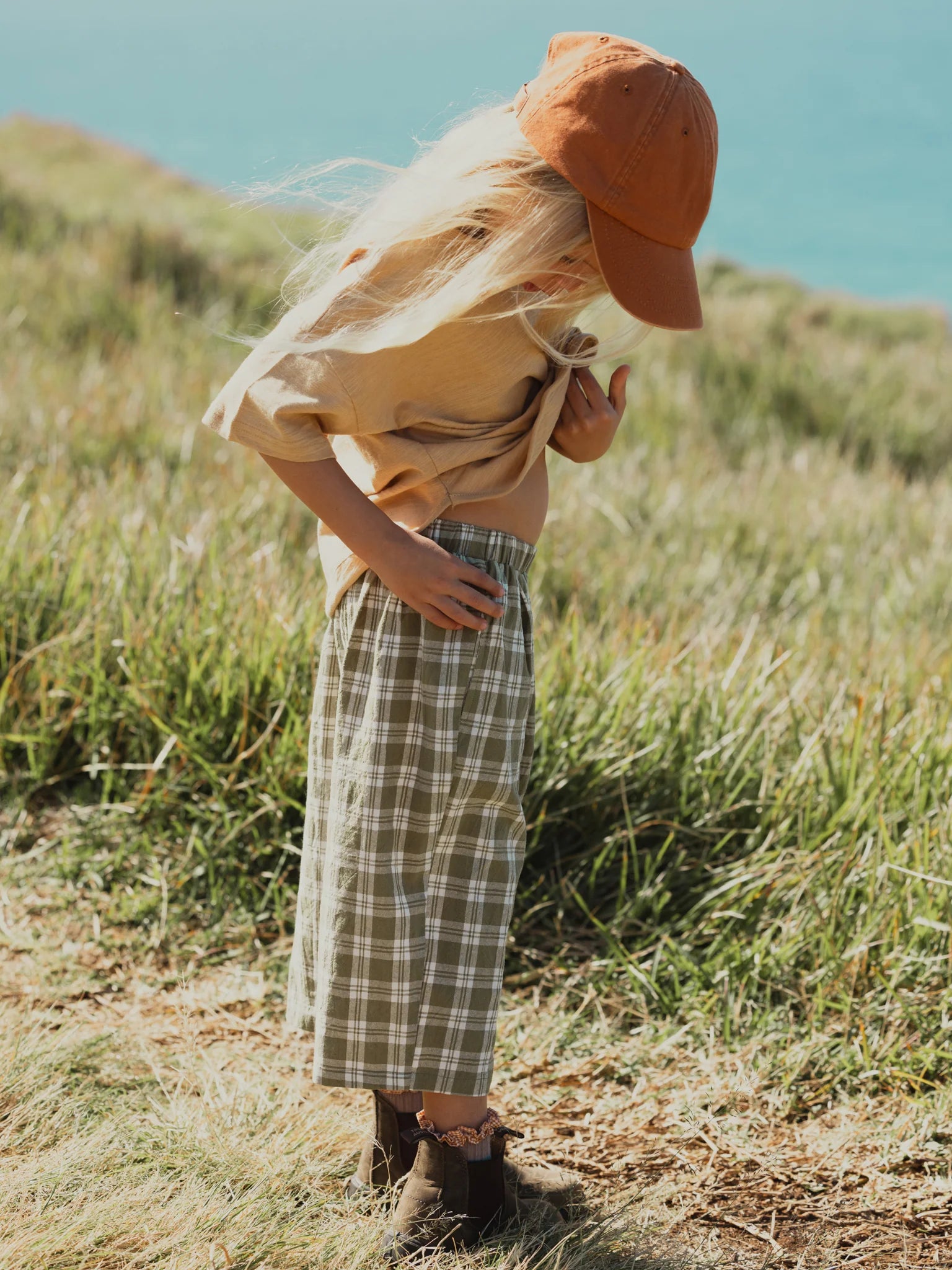 A child with long blond hair, wearing a brown cap, a beige shirt, VALENCIA BYRON BAY's Rio Pant in Olive (100% cotton plaid pants), and brown shoes, stands on grassy terrain near a body of water, looking down while slightly lifting their shirt. The sunlight illuminates the scene.