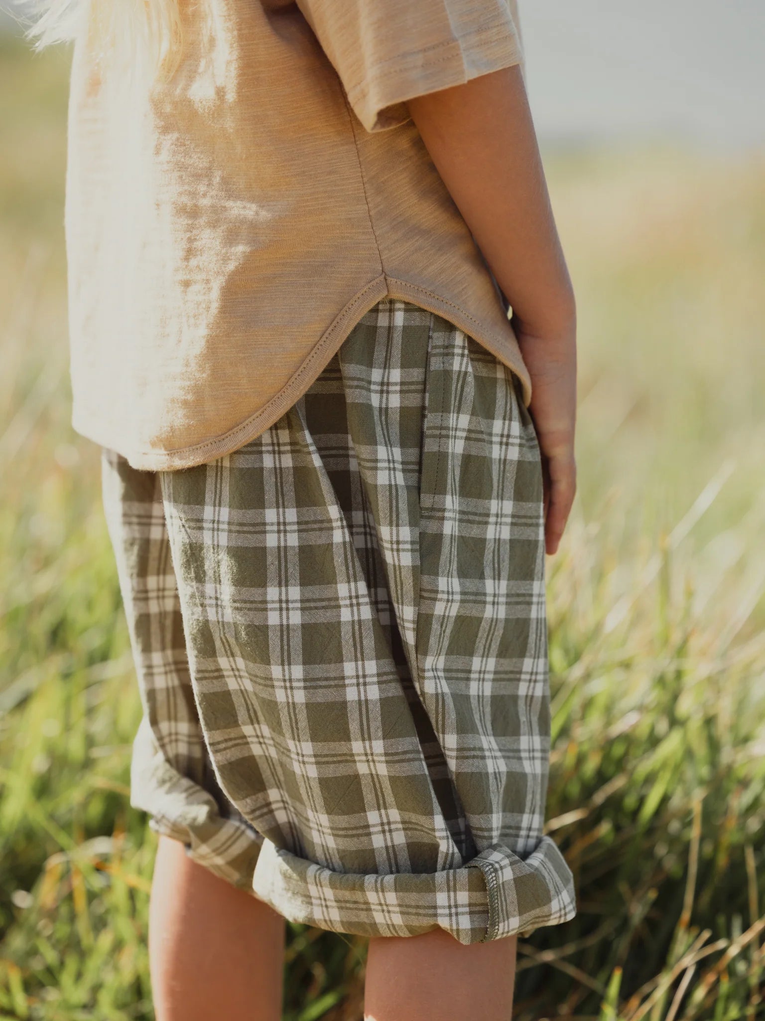 A child's lower torso and legs are shown, dressed in a light brown shirt and the VALENCIA BYRON BAY Rio Pant Olive with rolled-up cuffs, standing in a grassy field. The image showcases the unisex design and serene outdoor setting.